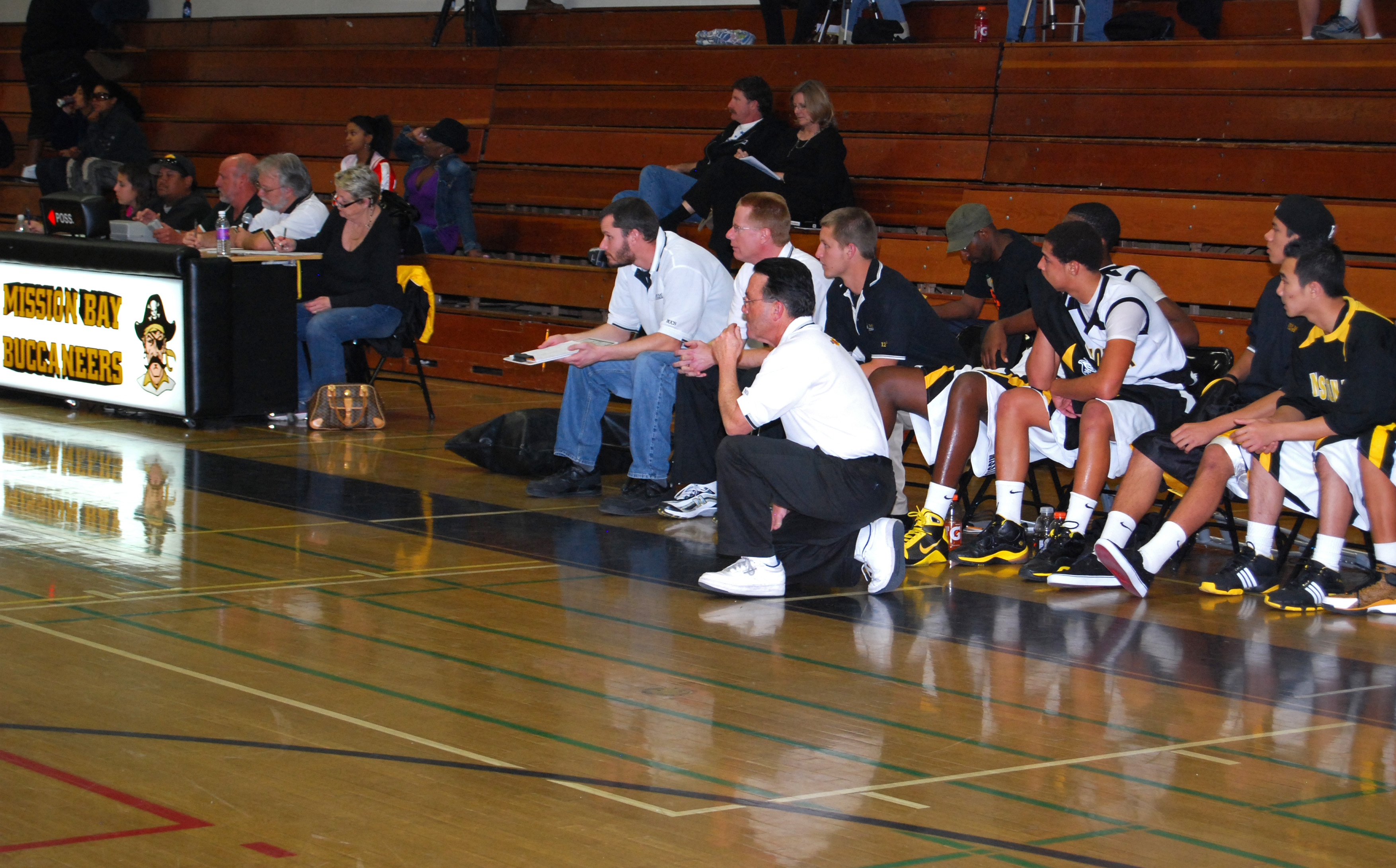 Coach Dennis Kane kneeling courtside directing his team from the bench during a game